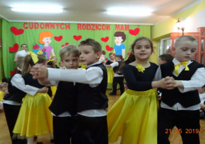 Dziewczynki w czarnych body i żółtych spódnicach oraz chłopcy ubrani na galowo tańczą w parach.